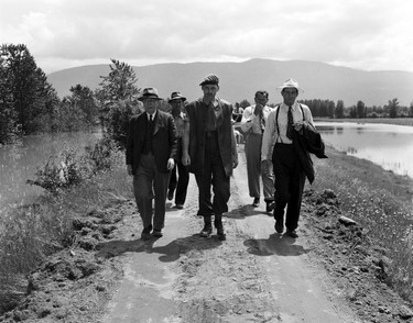 1948 Fraser River Flood - Sumas - Yarrows - A serviceman leads a group of men in suits down a dirt road flanked on both sides by water. 1948. Vancouver Sun.