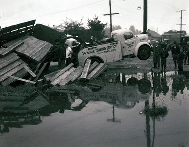 1948 Fraser River Flood - Trapp Rd. & BCE Barns - Specatators watch as the front end of a Royal City Motors tow truck is lifted into the air in its effort to help a flatbed truck out of flood waters. Wooden planks from the truck's bed can be seen toppling into the water. 1948. Vancouver Sun.