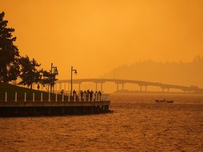 A person travels in a boat past people walking on the boardwalk as smoke from the McDougall Creek wildfire blankets the area on Okanagan Lake, in Kelowna on Aug. 18.