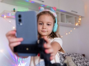 A little girl blogger wearing headphones records a video or story on her phone in her room against a background of color lights and flashlights. The content creator records vlog.