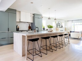 An open-plan kitchen-dining space stretches the length of the renovated home's main floor, with an oversized island offering seating for five, and a central sink. Oak veneer and sage green (Sherwin Williams Delft) cabinetry provide plenty of storage space, sleekly hiding a panelled fridge and built-in espresso station.