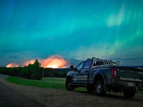 The wildfire forcing thousands to evacuate from a northeast British Columbia town has doubled again in size as the blaze grows merely a few kilometres west of city limits. The Aurora Borealis shines overhead of a B.C. Conservation Officer Service vehicle near the junction of highways 97 and 77, as a wildfire burns in the background near Fort Nelson, B.C., in a Saturday, May 11, 2024, handout photo.