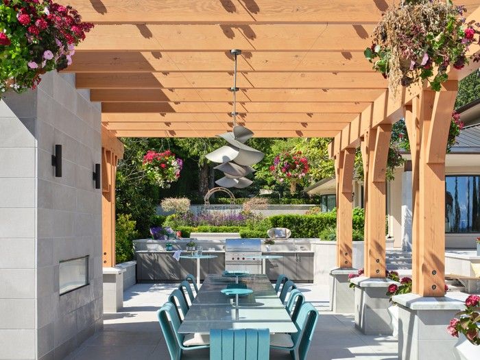  A glass-topped cedar pergola shelters an outdoor dining space with a built-in fireplace. A large glass dining table —Altopiano — Paola Lenti — pairs with ocean-inspired turquoise Frame chairs, also by Paola Lenti (sourced by Livingspace Vancouver), under soft-lighting pendant lights (Hubbardton Forge Hibiscus Pendants).