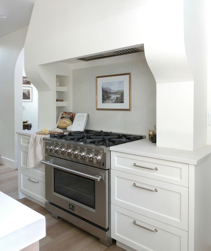  A curvy custom-framed hood fan houses built-in shelves, while a quartz slab backslash is grout-free and easy to clean, leaving space for an unexpectedly placed piece of artwork over the range.
