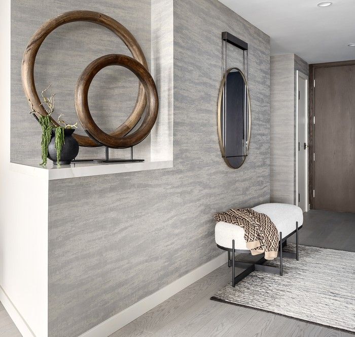  A wide entry sets the visual tone for the home, with grey-washed flooring complementing Colour & Design wallpaper from Metro Wallcoverings installed by Revive Wallcoverings. Arteriors mirrors, ring sculptures and vases style a built-in niche, while a CF Interiors bench adds seating.