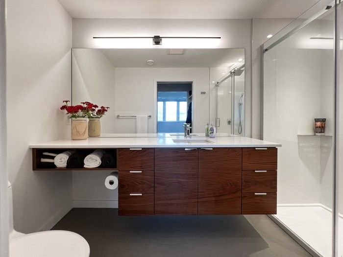 The bathroom carries the considered balance of function and light. A floating walnut vanity with open shelving elevates the compact space, complemented by a wide mirror and slim light fixture.  The bathroom carries the considered balance of function and light. A floating walnut vanity with open shelving elevates the compact space, complemented by a wide mirror and slim light fixture.