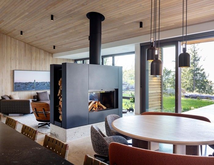 In the main cabin, a two-sided wood-burning fireplace by Stûv divides the living and dining areas without interrupting flow. Mid-century modern-inspired furniture and lighting contrasts the more rustic feel of the woods. Curved Sendai seating by Minotti and a custom dining table by Vancouver furnituremaker Lock & Mortise bring a soft counterpoint to the cabin's clean-lined architecture, under blackened brass Aria pendants by Allied Maker.
