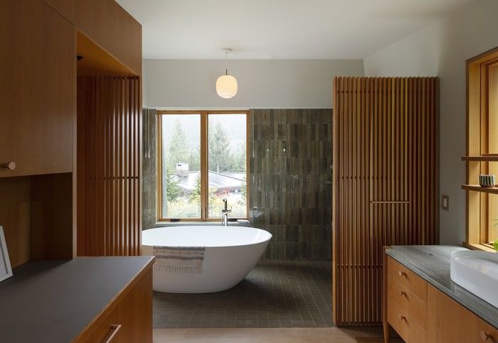 Brown-green Italian porcelain tile (Lume by Marazzi) wraps a bathroom and wet area in the ensuite, while a bathtub sits beside a sloped window that follows an exterior wall and opens a view toward the mountains. A Louis Poulsen VL 45 Radiohus Pendant hangs overhead.