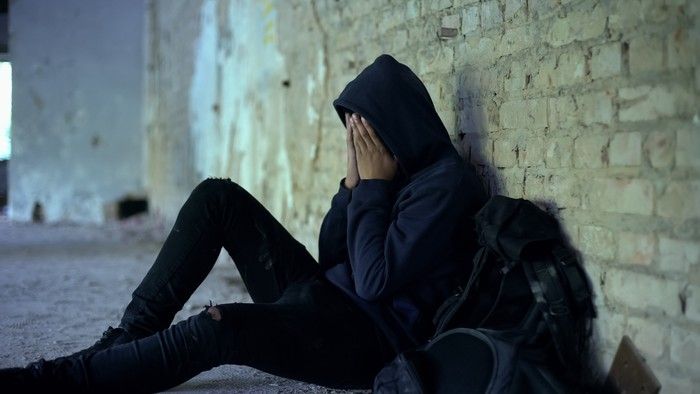 A teenager wearing a hoodie sits with his hands over his face.