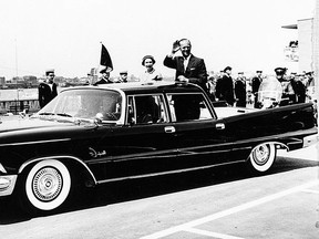 Standing next to Queen Elizabeth II, Prince Philip waves to crowds at the docks from the back of a Chrysler Imperial.  At left are crew members of the Royal Yacht Britannia.