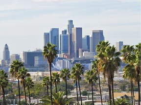 The Los Angeles skyline is seen through a row of palm trees on a partly cloudy day.