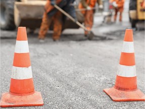 Road workers repair a road with shovels and asphalt, with orange cones in the foreground.