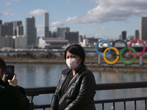 In this Jan. 29, 2020, photo, a tourist wearing a mask poses for a photo with the Olympic rings in the background, at Tokyo's Odaiba district.