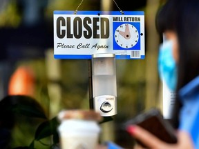In this file photo a pedestrian wearing her facemask and holding a cup of coffee walks past a closed sign hanging on the door of a small business.