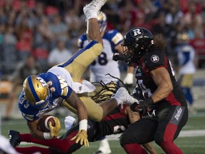 Ottawa Redblacks defensive lineman J.R. Tavai, right, looks on as Winnipeg Blue Bombers running back Andrew Harris, left, is upended by defensive back Sherrod Baltimore during second quarter CFL action in Ottawa, Friday.