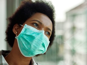 Distraught black woman with face mask thinking while looking through the window at home.