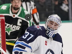 The puck deflects off of Winnipeg Jets goaltender Connor Hellebuyck as Dallas Stars left winger Jamie Benn looks on Friday night in Dallas. The Stars would later score a contentious goal on Hellebuyck after Benn bulldozed him and ripped the goalie mask clear off the netminder’s head. The Jets went on to win 5-4 in overtime.